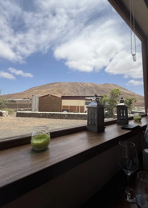 Vista desde la mesa a través del ventanal de Casa Marcos: faroles, vela verde, copas y una montaña volcánica bajo cielo azul